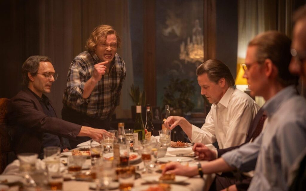 A dinner with four characters. One of them standing and pointing at the others.
